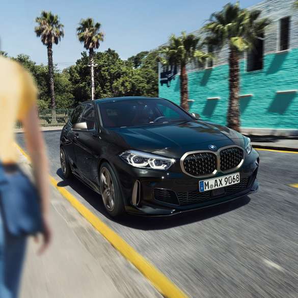 BMW M135i xDrive, three-quarter front view. BMW M135i xDrive in Black Sapphire metallic, three-quarter front view driving, with woman in foreground.