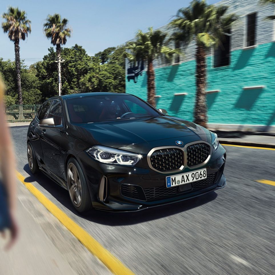 BMW M135i xDrive, three-quarter front view. BMW M135i xDrive in Black Sapphire metallic, three-quarter front view driving, with woman in foreground.
