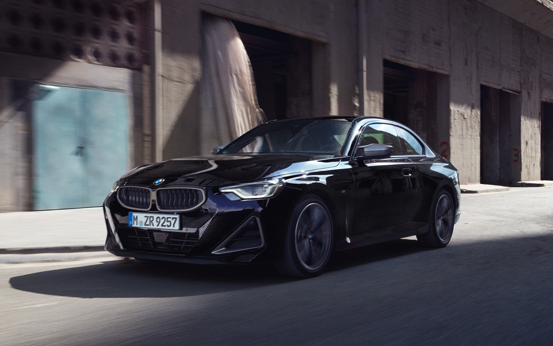 BMW M240i xDrive Coupé G42 2021 Black Sapphire metallic three-quarter side view driving in front of warehouses